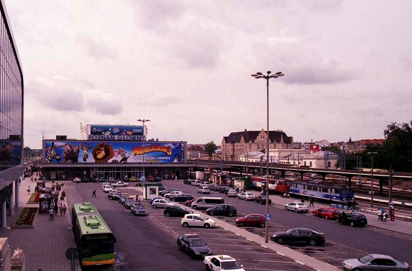 widok na budyek starego dworca PKP, przed dworcem plac, na którym zatrzymują się samochody i autobusy, na fasadzie dworca duża reklama - grafika artykułu