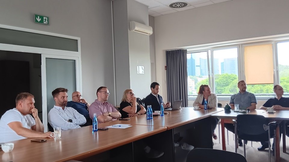 grupa ludzi siedzi przy stole konferencyjnym, uważnie na coś patrzą i słuchają