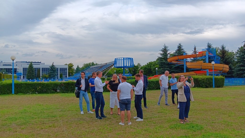 grupa ludzi na trawniku, rozmawiają, rozglądają się, wokół znajdują się urządzenia rekreacyjne i sportowe, na przykład zjeżdżalnia do basenu