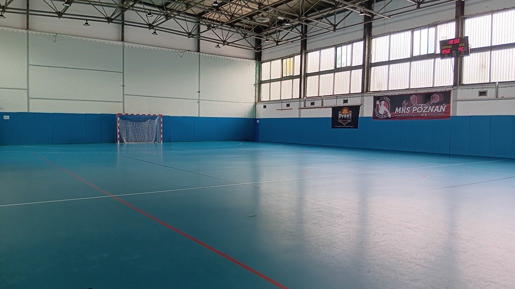 Hala sportowa, pusta, nawierzchnia w kolorze niebieskim, widoczna jedna bramka do piłki ręcznej i tablica świetlna