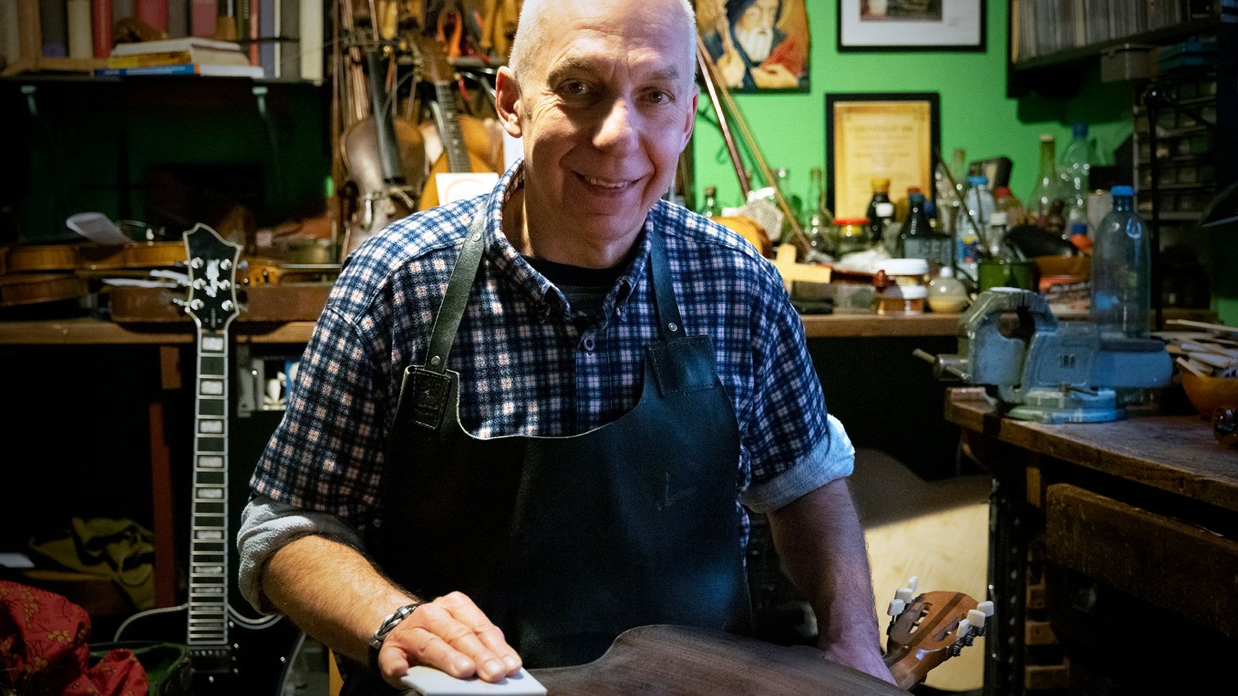 Benedykt Niewczyk - tytuł Zasłużony dla Miasta Poznania 2025 - mężczyzna w wieku senioralnym siedzi i uśmiecha się ma kraciastą koszulę i fartuch, znajduje się w pracowni lutniczej, za nim widoczne są skrzypce i gitara