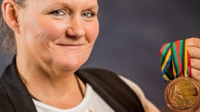 Izabela Dylewska-Światowiak - tytuł Zasłużony dla Miasta Poznania 2025 - kobieta w średnim wieku, włosy spiętedo tyłu, uśmiecha się, w ręku trzyma medal na kolorowej szarfie