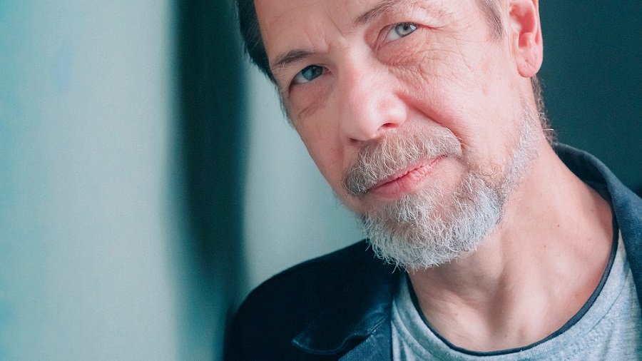 Przemysław Czapliński - Nagroda Naukowa Miasta Poznania 2025 - mężczyzna w średnim wieku, włosy zaczesane na bok, zarost na twarzy, włosy siwiejące, opiera się o ścianę, głowa lekko przechylona