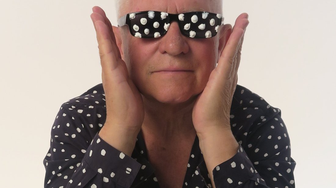 Piotr C. Kowalski - Nagroda Artystyczna Miasta Poznania 2025 - starszy człowiek, siwe włosy, ubrany w czarną koszulę w białe kropki i ciemne okulary z białymi kropkami, głowę podpiera dłońmi