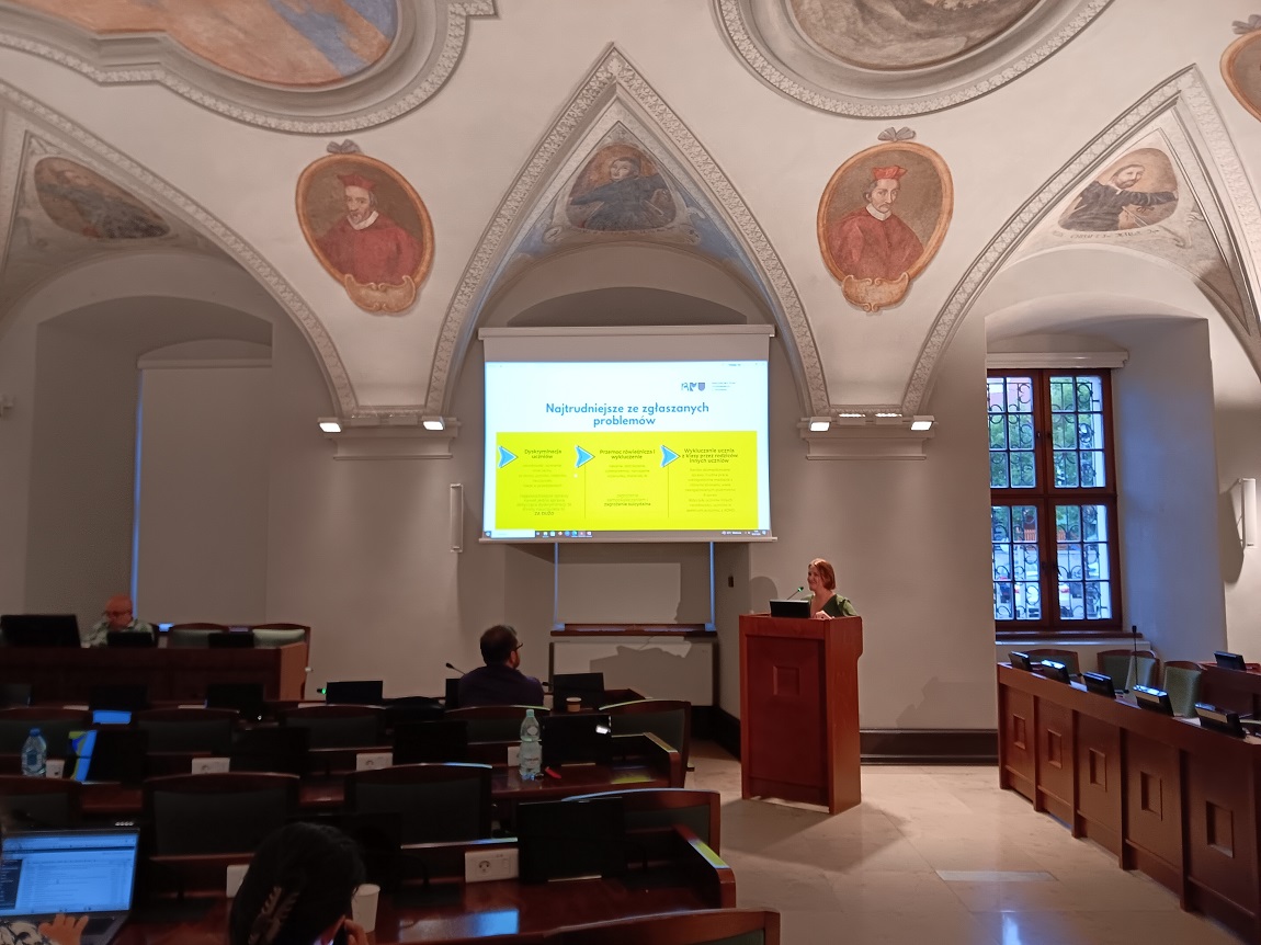 sala obrad z sufitem zdobionym kolorowymi freskami, jedna osoba stoi przy mównicy, na ekranie wyświetlona jest prezentacje, pozostałe osoby siedzą i słuchają - grafika artykułu