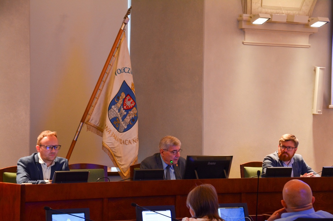 trzy osoby siedzą przy stole prezydialnym w sali, za nimi sztrandar z herbem Poznania - grafika artykułu