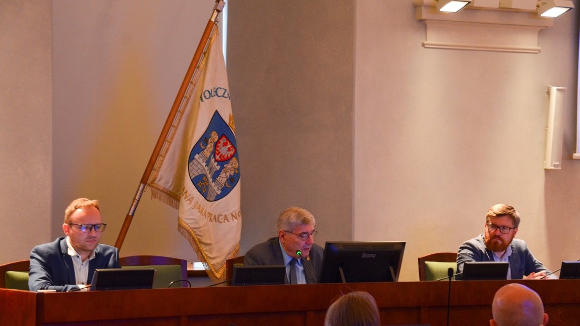 trzy osoby siedzą przy stole prezydialnym w sali, za nimi sztrandar z herbem Poznania