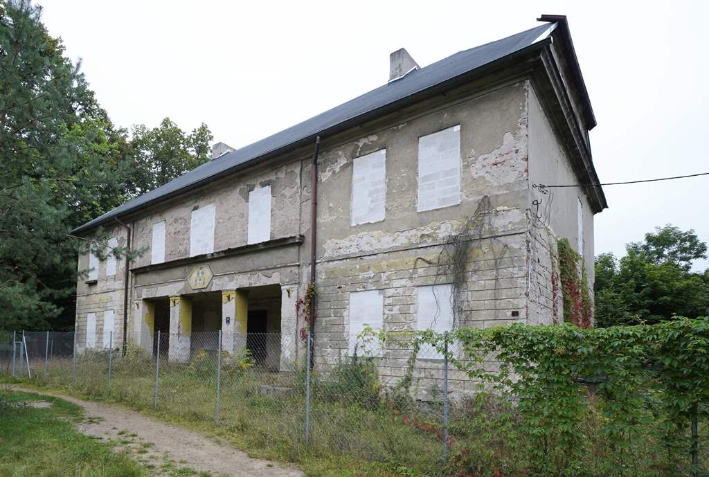 budynek dwukondygnacyjny ze spadzistym dachem, w otoczebniu drzew i trawy, ogrodzony płotem siatką, ma zamurowane okna, fasada z odpadającym tynkiem - grafika artykułu