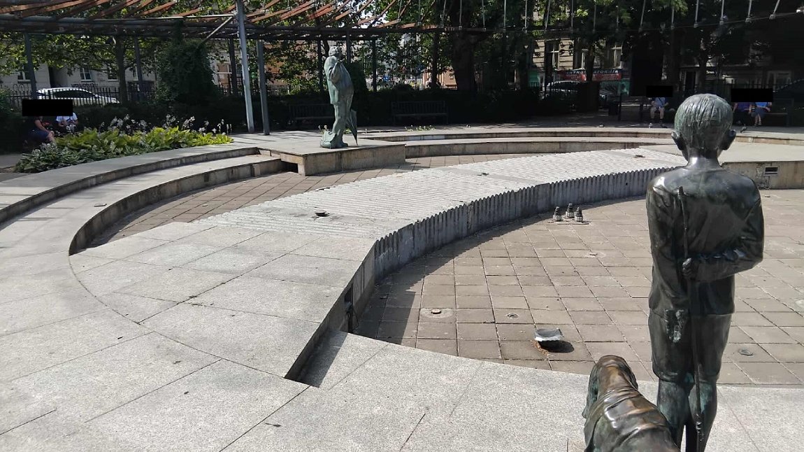 skwer otoczony zielenią, niedziałąjąca fontanna, jej elemantami są rzeźby starszego mężczyzny i chłpca z psem