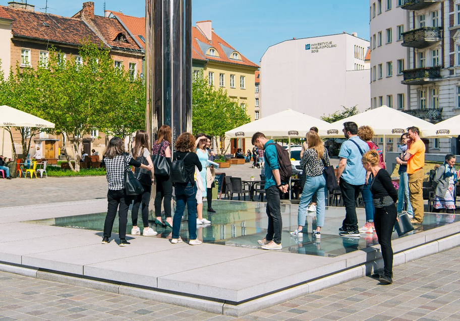 plac z ogródkami gastronomicznymi, wokół kamienice, w posadzce placu ekspozycja za szklanymi szybami, ogląda ją grupa osób patrząc w dół, nad espopzycją wznosi się stella z lustrzaną powiechrznią - grafika artykułu