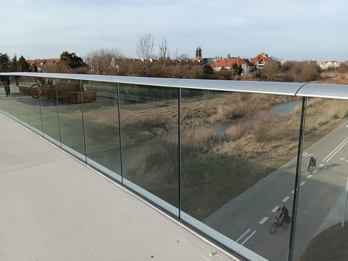 fragment przeszklonej balustrady mostu pieszo rowerowego, za nią widok na ścieżkę rowerową i tereny nad rzeką - grafika artykułu