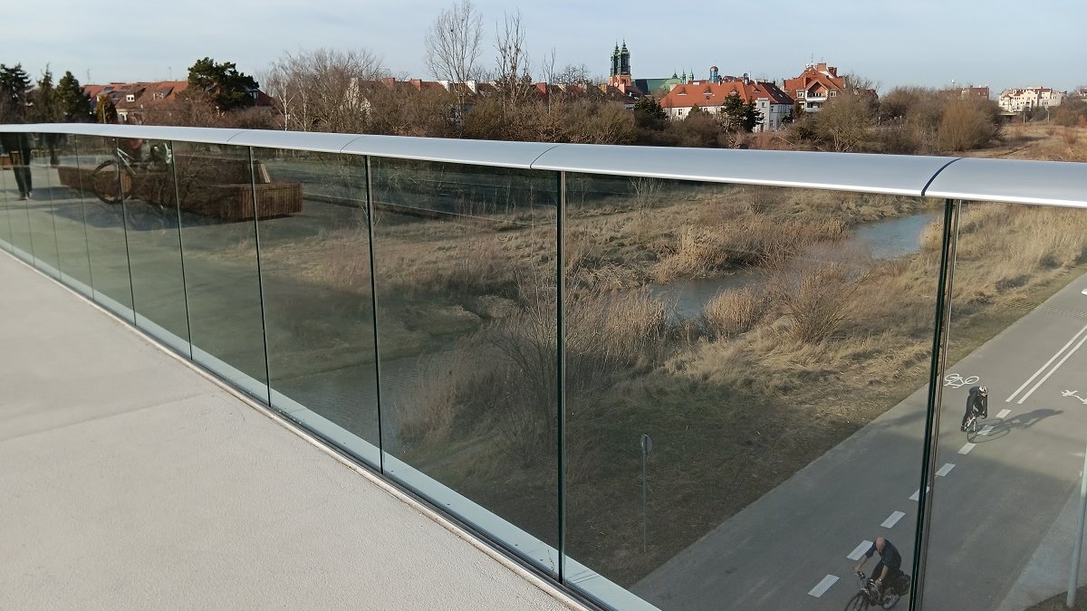 fragment przeszklonej balustrady mostu pieszo rowerowego, za nią widok na ścieżkę rowerową i tereny nad rzeką