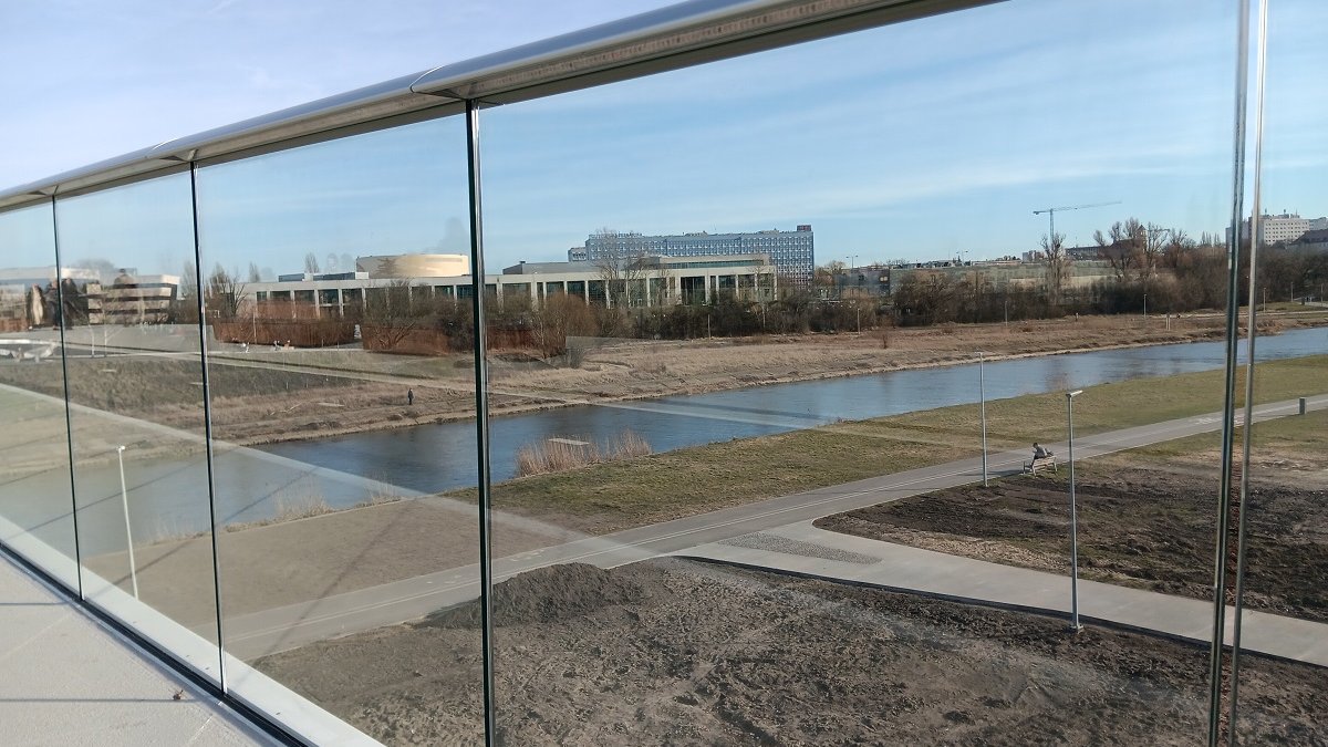 fragment przeszklonej balustrady mostu pieszo rowerowego, za nią widok na ścieżkę rowerową i tereny nad rzeką