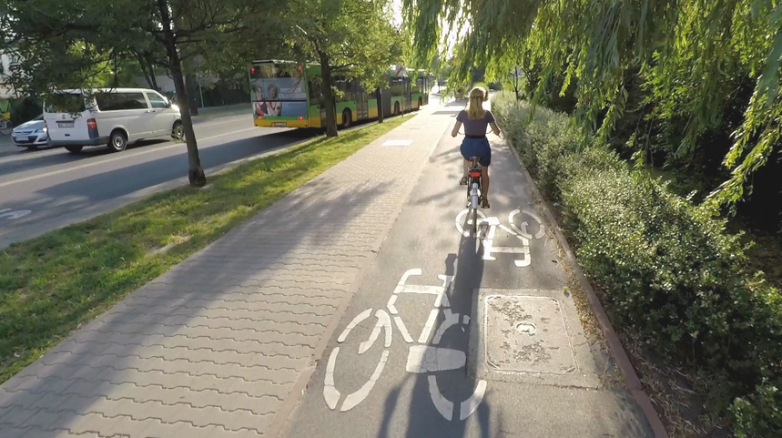 droga rowerowa i chodnik, na drodze rowerowej jedzie rowerzystka, po obu stronach zieleń - drzewa i krzewy, na ulicy jadą dwa samochody i autobus komunikacji miejskiej - grafika artykułu