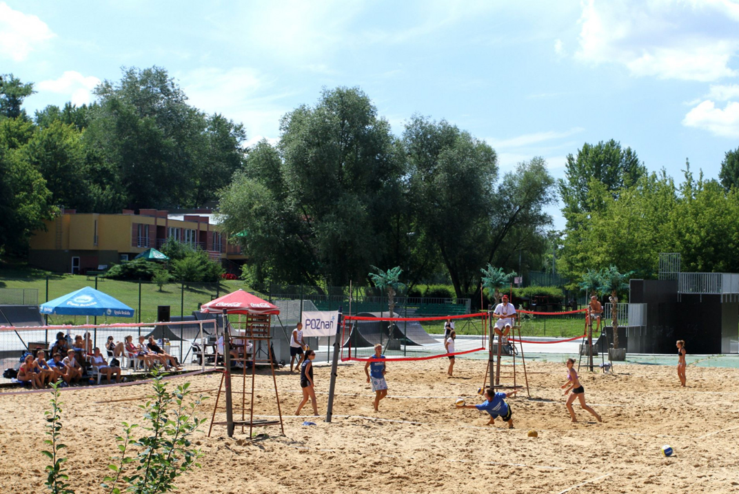kilka osób gra w siatkówkę plażową, inne osoby oglądają mecz, jest słoneczna pogoda, za boiskiem plażowym widać skatepark i dalej drzewa - grafika artykułu
