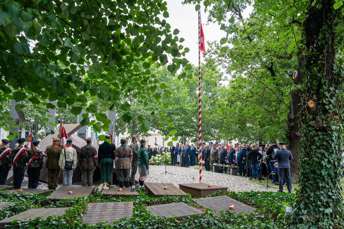na placu przed pomnikiem odbywają sie uroczości z udziałem harcerzy i oficielu, na placu masz z biało czerwoną flagą, plac otaczają drzewa - grafika artykułu