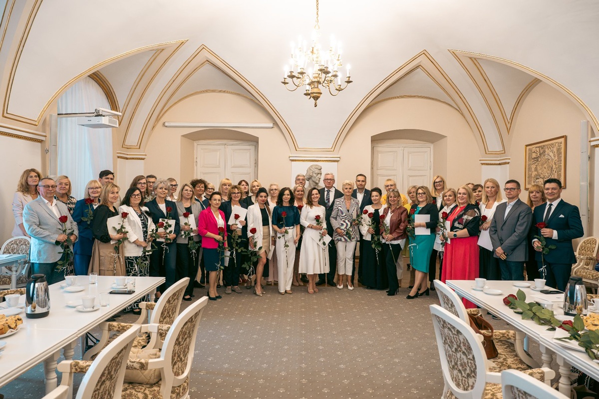 grupa około kilkudziesięciu osób kobiet i mężczyzn pozuje do zdjęcia w zabytkowej reprezentacyjnej, jasnej sali, są ubrani elegancko, uśmiechnięci, niektórzy trzymają kwiaty - grafika artykułu
