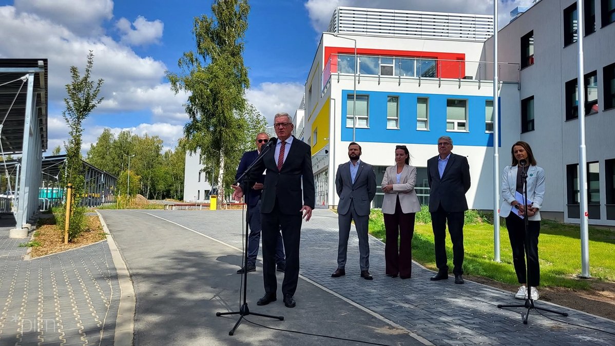 kilka osób stoi w rzędzie, jedna przy mikrofonie na stojaku, stoją przed budynkiem szkoły