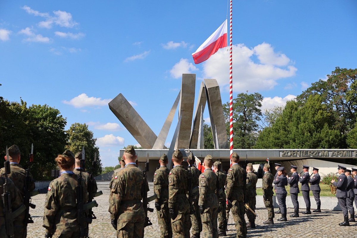 grupa żołnierzy stoi na placu przed pomnikiem, jest maszt a na nim flaga biało czerwona - grafika artykułu