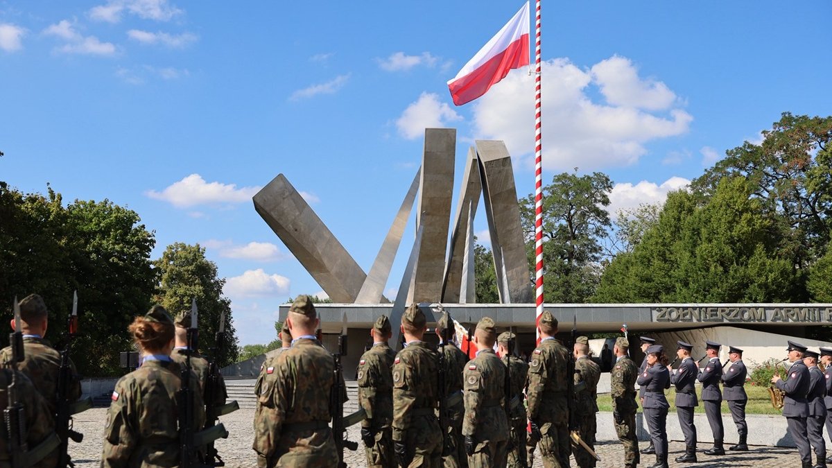 grupa żołnierzy stoi na placu przed pomnikiem, jest maszt a na nim flaga biało czerwona