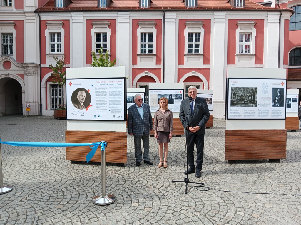 Otwarcie wystawy poświęconej Cyrylowi Ratajskiemu trzy osoby stoja na tle plansz wystawowych, jedna z nich przemawia do mikrofonu - grafika artykułu