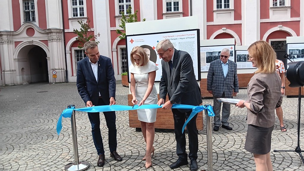 Otwarcie wystawy poświęconej Cyrylowi Ratajskiemu - kilka osób przecina symboliczna wstęgę, za nimi widoczne plansze wystawowe