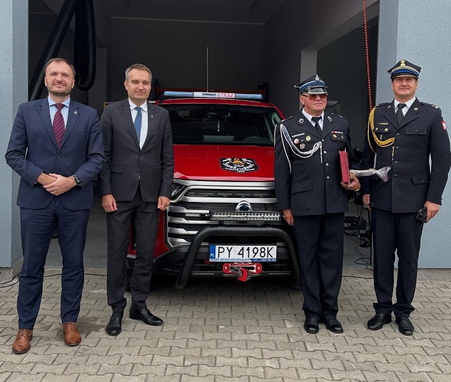 czterwch mężczyzn dwóch w garnirurach i dwóch w galowych mundurach strażaków pozuje do zdjęcia na tle terenowego samochodu strażackiego - grafika artykułu