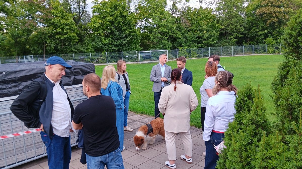 grupa osób stoi przed boiskiem trawiastym, rozmawiają, jedna z osób trzyma psa na smyczy