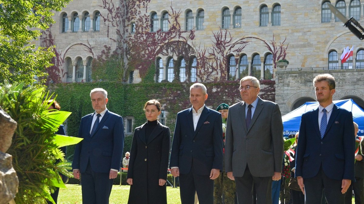 kilka osób ubranych na galowo stoi w parku przed pomnikiem składając wiązanki kwiatów