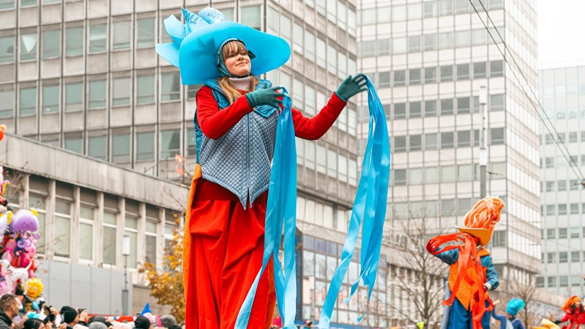 korowód idący przez ulicę widoczni ludzie w barwnych strojach na podwyższeniu wzdłuż ulicy utawieni ludzie oglądający przemarsz korowodu