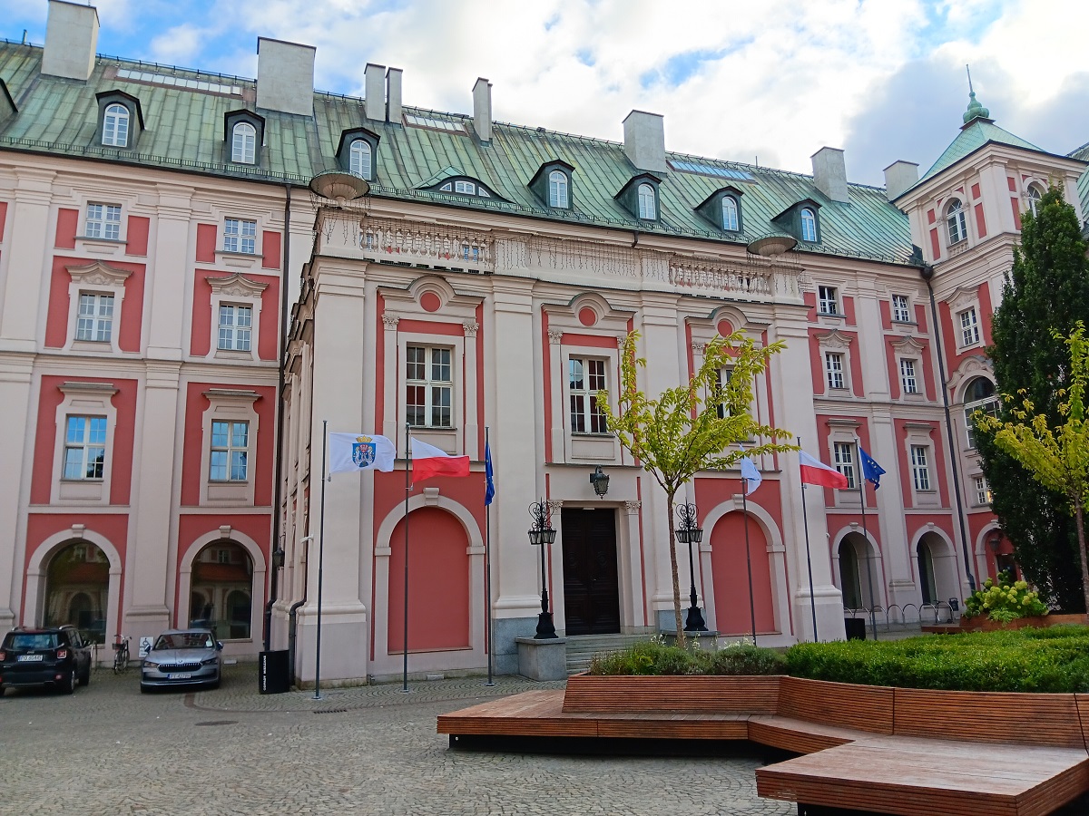gmach od strony dziedzińca zabytkowa architektura przed wejście do gmachu ławki i zieleń oraz flagi na masztach flaga Poznania Polski i Unii Europejskiej - grafika artykułu