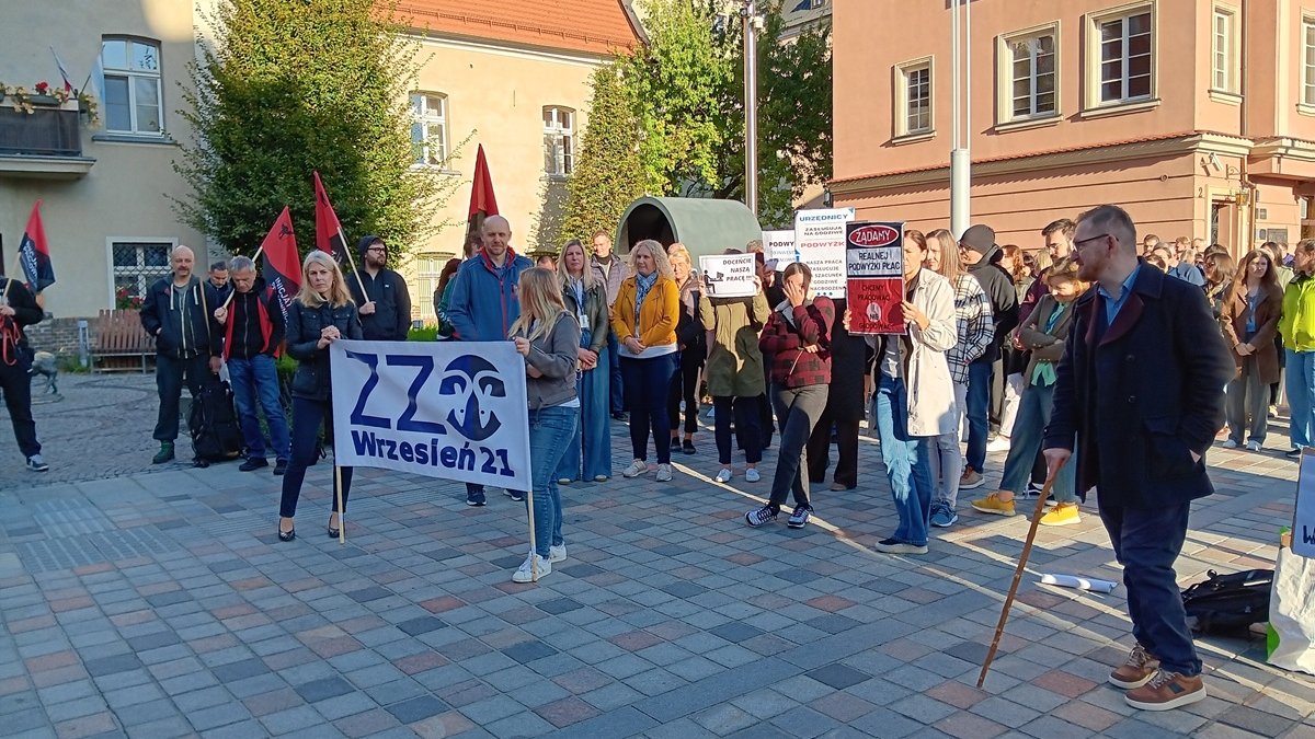 grupca około kilkudzisięciu osób stoi na placu z transparentami i flagami wokół placu budynki