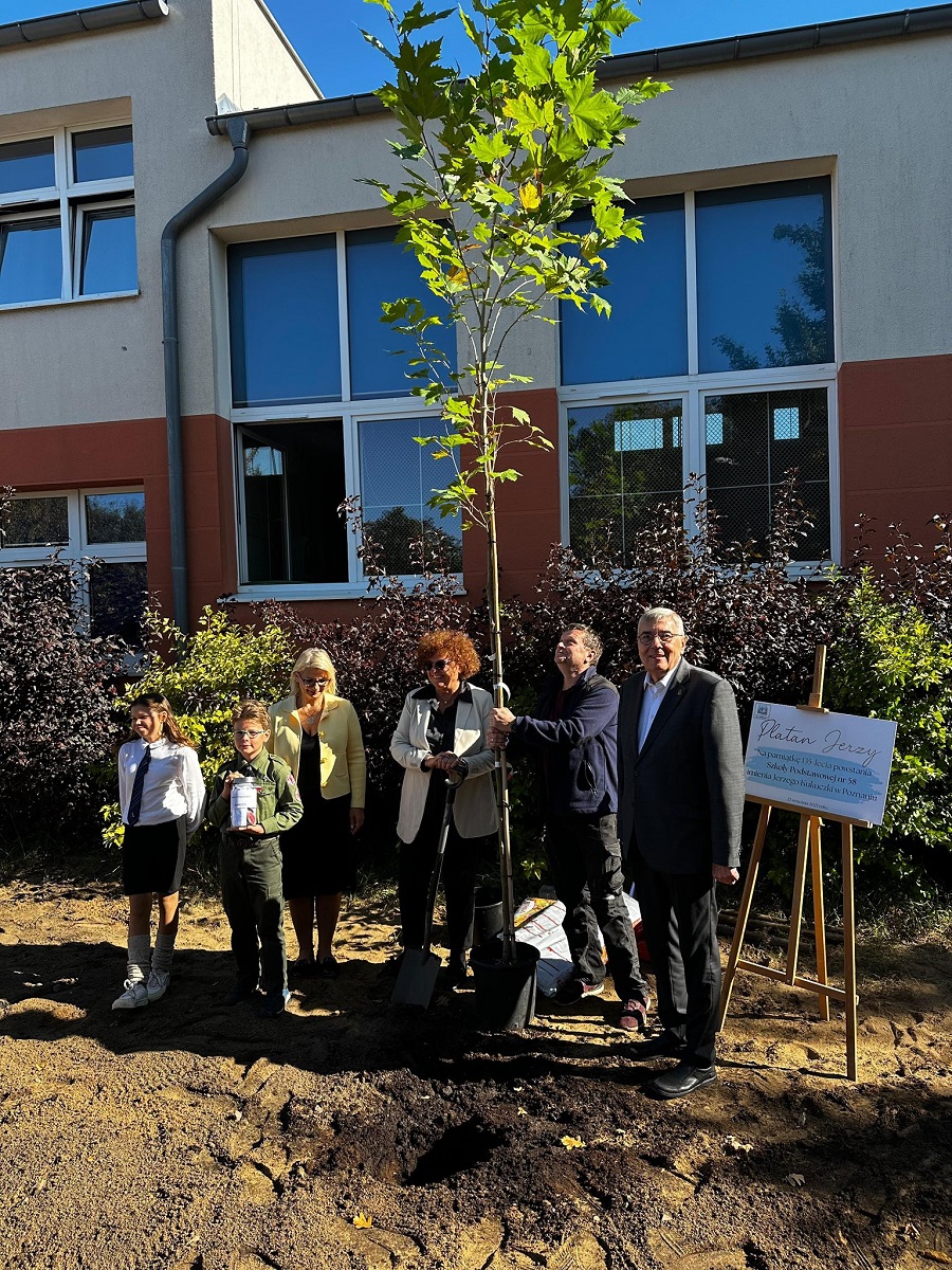grupa osób pozuje do zdjęcia przy nowo posadzonym drzewie przed budynkiem szkolnym - grafika artykułu