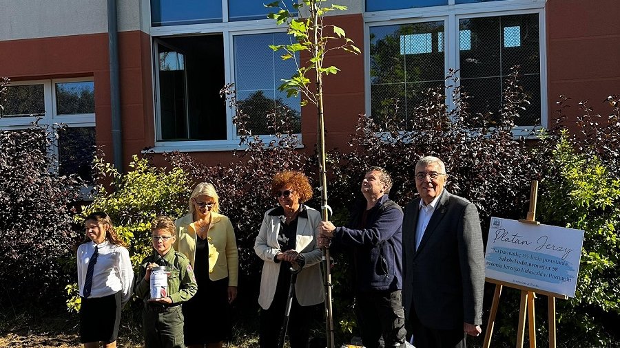 grupa osób pozuje do zdjęcia przy nowo posadzonym drzewie przed budynkiem szkolnym