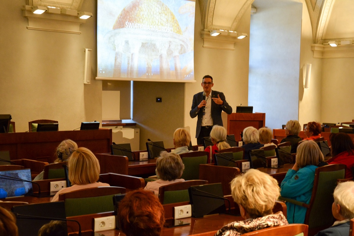 w dużej sali grupa osób siedzi przy stanowiskach z mikrofonami przed sto nimi mężczyzna z mikrofonem i mówi do nich - grafika artykułu