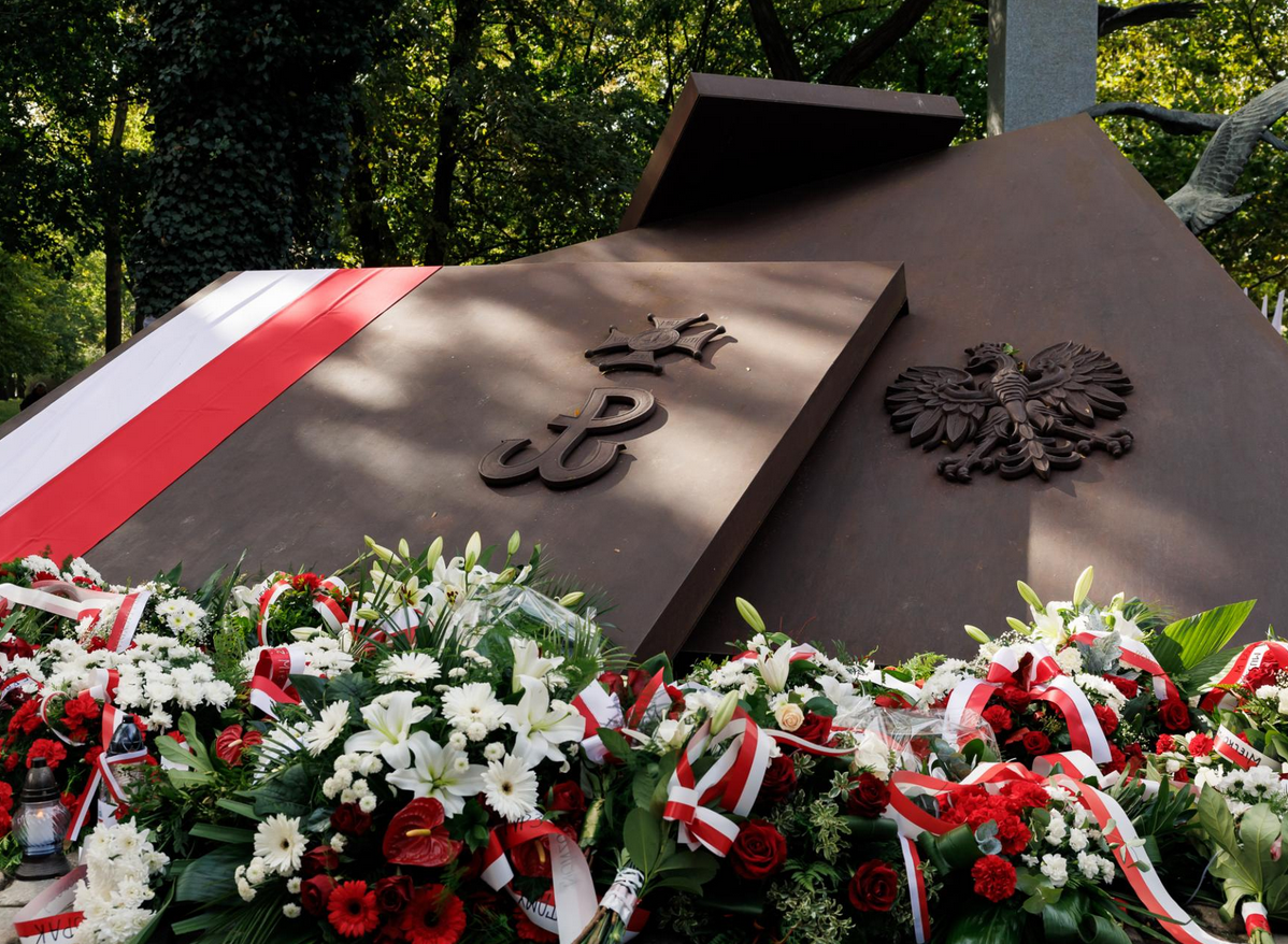 pomnik w kształcie nałożonych na siebie dużych płyt na nich wizerunki godła Polski symbolu harcerstwa i Polski Walczącej pod pomnikiem złożone wiązanki kwiatów i biało czerwona flaga - grafika artykułu