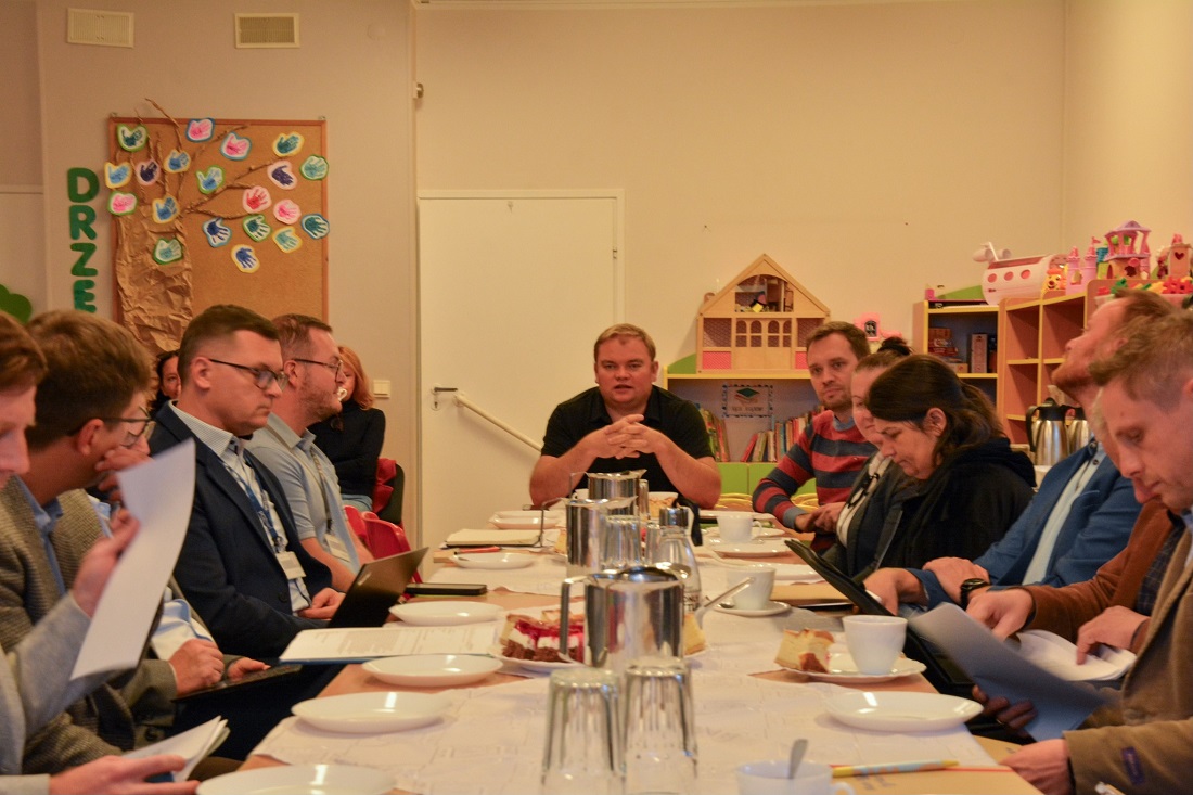 grupa osób kobiet i mężczyzn siedzi przy podłużnym stole, prowadzą spotkanie, na stole dokumenty, laptopy, termosy do napojów, taleczyki i filiżanki - grafika artykułu