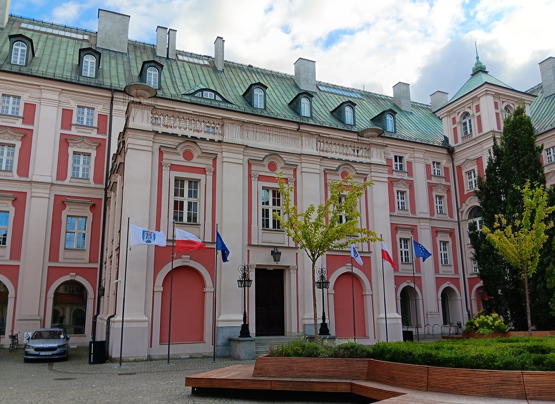 gmach Urzędu Miasta Poznania od strony dziedzińca zabytkowa architektura przed wejście do gmachu ławki i zieleń oraz flagi na masztach flaga Poznania Polski i Unii Europejskiej - grafika artykułu