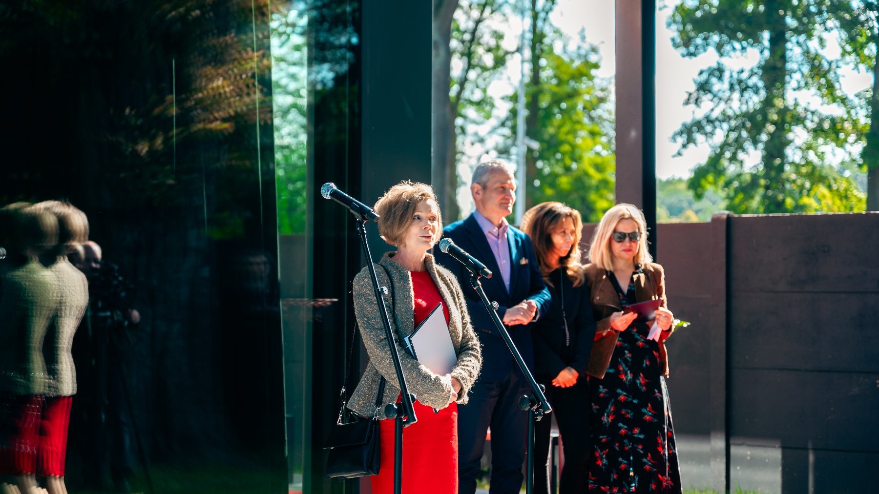 przed budynkiem z dużymi panoramicznymi oknami stoją trzy kobiety i mężczyzna jedna z kobiet stoi przy mikrofonie ze statywem