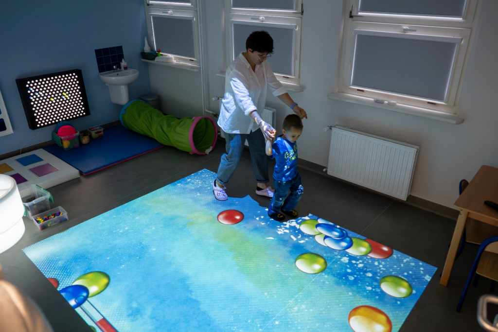 Sala dla terapii dzieci dziecko przy pomocy opiekunki korzysta z urządzenia wyświetlającego w podłodze barwne obrazy - grafika artykułu