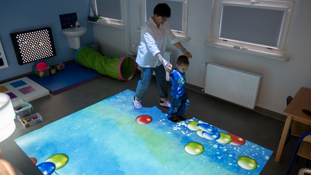 Sala dla terapii dzieci dziecko przy pomocy opiekunki korzysta z urządzenia wyświetlającego w podłodze barwne obrazy