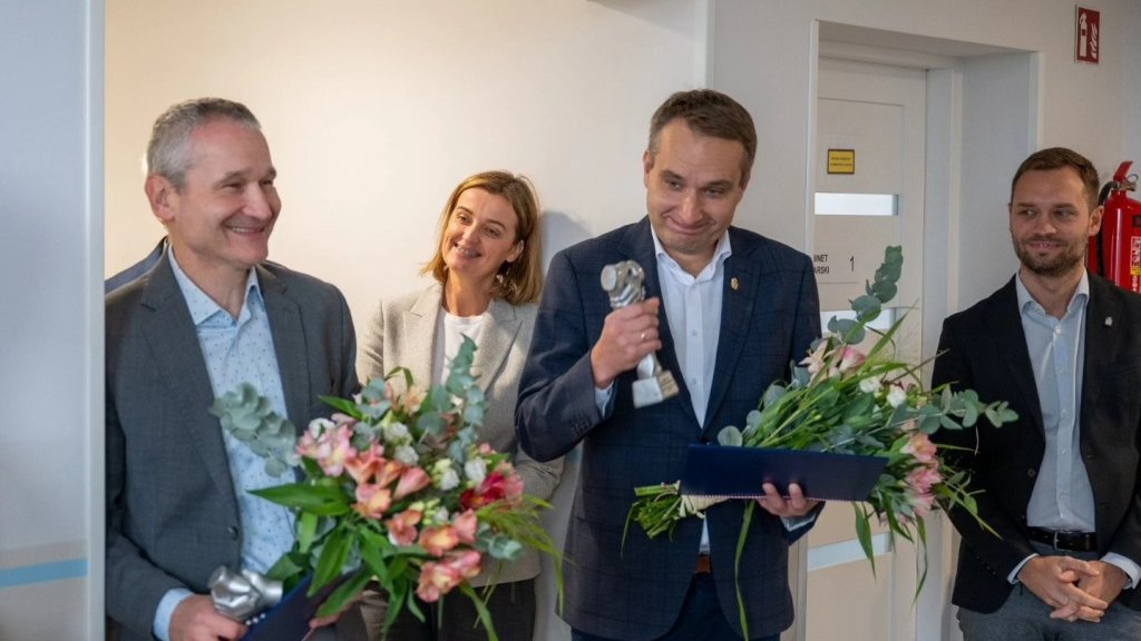 trzech mężczyzn w marynarkach za nimi kobieta dwóch z mężczyzn trzyma bukiety kwiatów a jeden z nich dodatkowo statuatkę