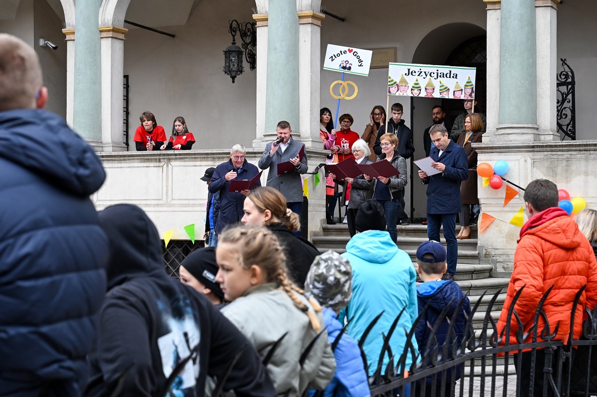 na schodach przed ratuszem grupa kilku osób kobiet i mężczyzn część z nich czyta książkę za nimi osoby z transparentami Złote Gody i Jeżycjada przed nimi zgromadzona młodzież i dzieci - grafika artykułu