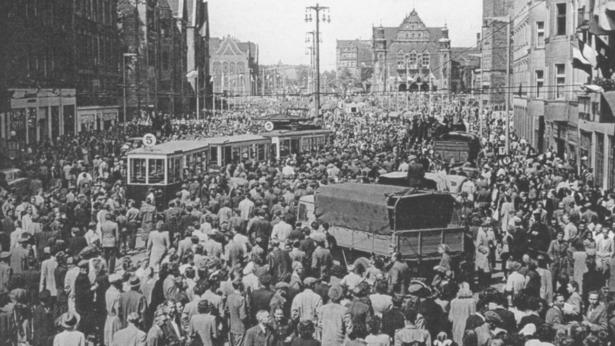 czarno białe archiwalne zdjęcie przestawiające tłum demontrantów w zabudowie śromiejskiej w tłumie samochodwy ciężarowe i tramwaje