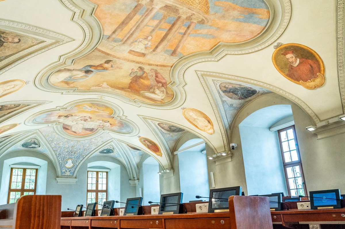 sala sesyjna Rady Miasta Poznania widać sufit zdobiony barwnymi zabytkowymi freskami oraz stanowiska dla radnych z monitorami i mikrofonami - grafika artykułu