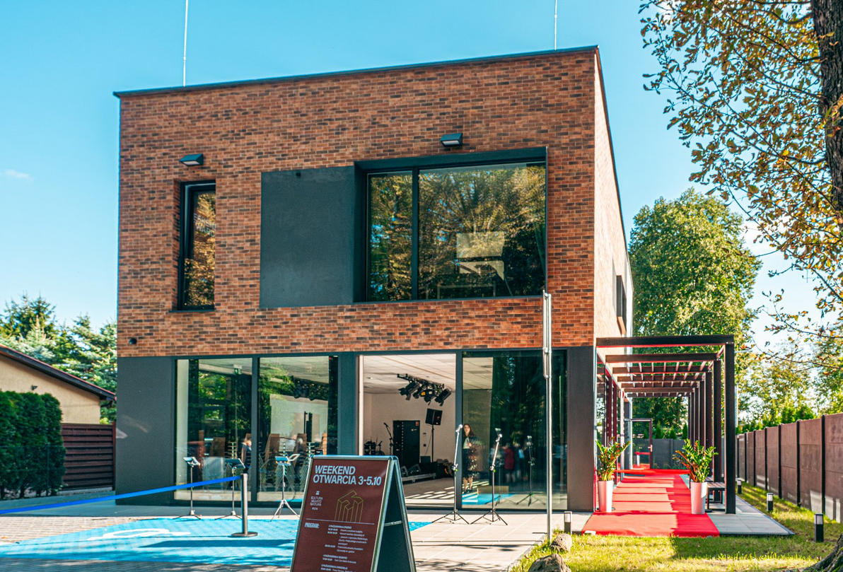 nowoczesny, dwupiętrowy budynek o elewacji z ciemnej cegły i dużymi, panoramicznymi oknami, otwarty na czerwony dywan prowadzący na zewnątrz. Przed wejściem stoi tablica informacyjna z napisem "WEEKEND OTWARCIA 3-5.10", a budynek jest otoczony zielenią - grafika artykułu - grafika artykułu