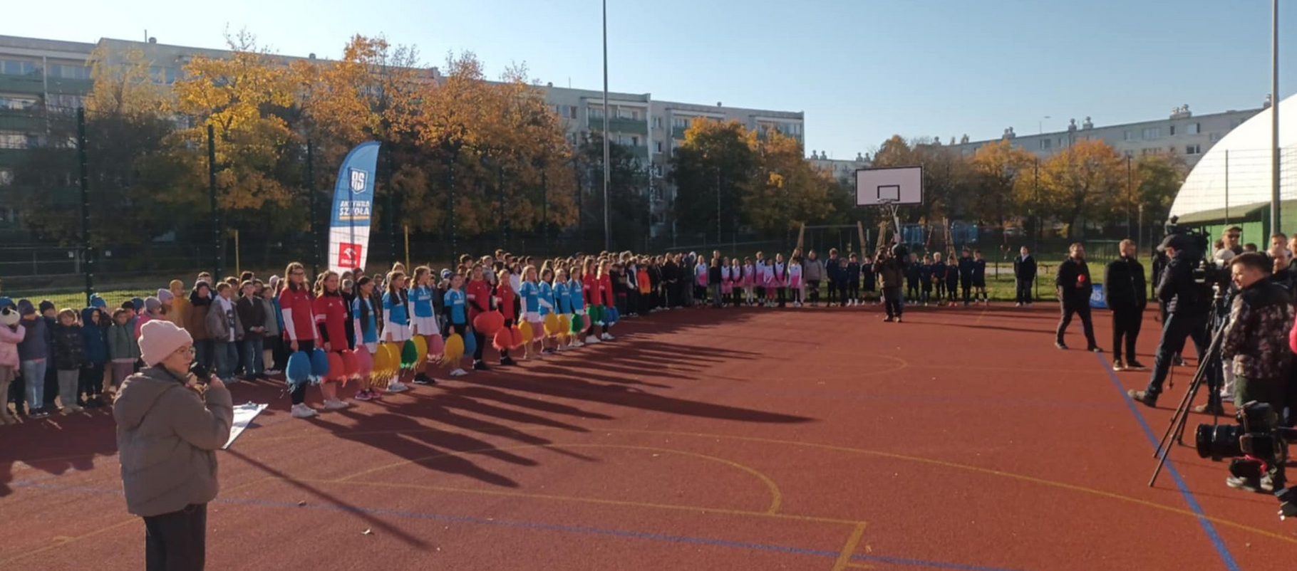 boisko szkolne na nim duża grupa dzieci i młodzieży poza boiskiem niskie bloki i drzewa - grafika artykułu