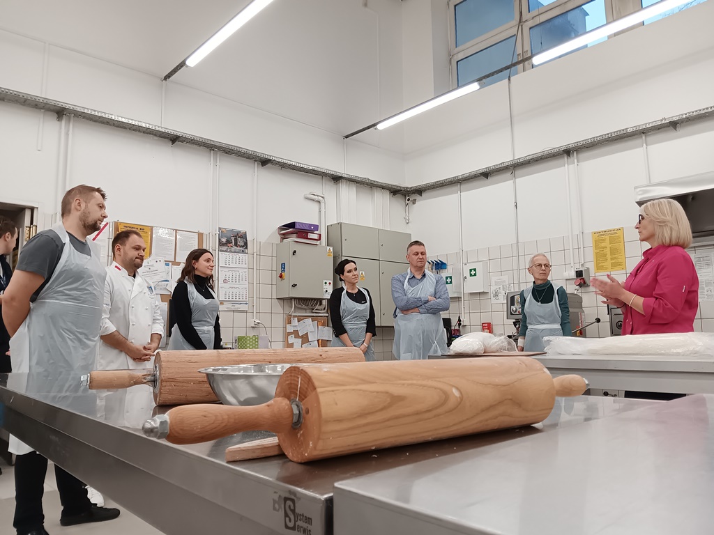 Grupa osób w fartuchach w sali piekarniczej przed nimi stół na nim dwa wałki do ciasta - grafika artykułu