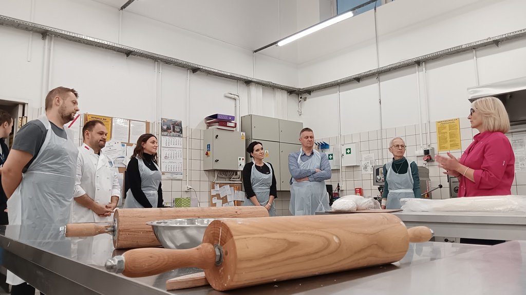 Grupa osób w fartuchach w sali piekarniczej przed nimi stół na nim dwa wałki do ciasta