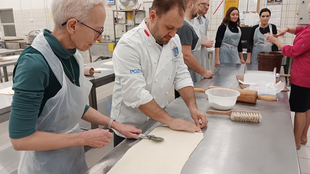 mężczyzna pokazuje kobiecie jak kroi się ciasto za nimi inna kobieta pokazuje grupie osób jak nakłada się masę do worka piekarniczego